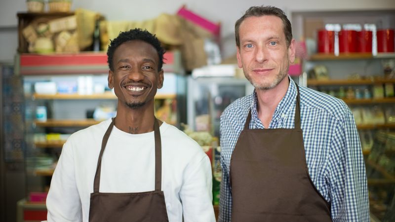 Dois donos de um comércio de bairro sorrindo atrás do balcão da loja